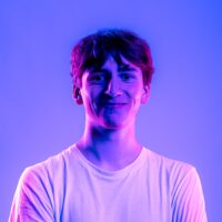 Ein junger Mann mit dunklen, lockigen Haaren und weißem T-Shirt lächelt vor einem blau-violetten Hintergrund